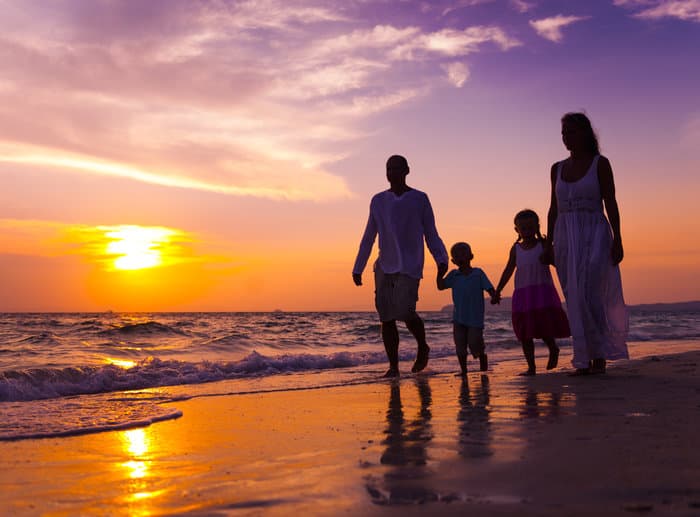 A silhouetted family walks hand-in-hand along the beach at sunset.