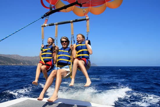 Three people, two children and an adult, are parasailing over the ocean, all wearing life jackets and smiling.