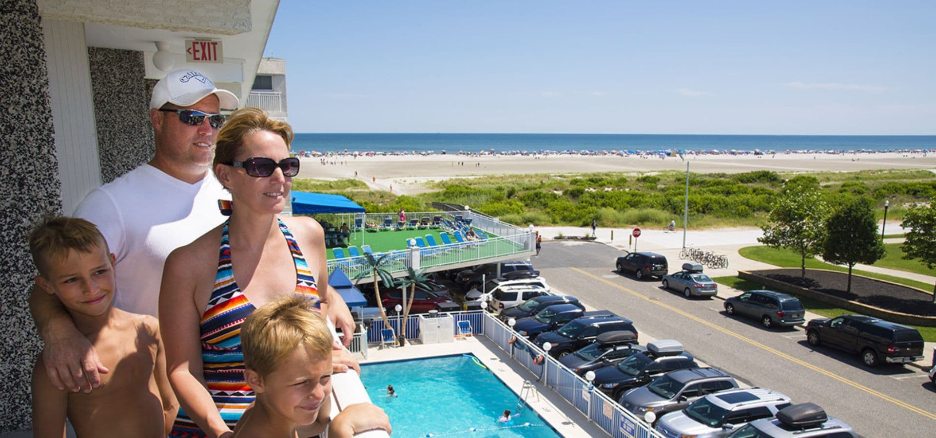 A family stands on a balcony overlooking a pool and beach on a sunny day.