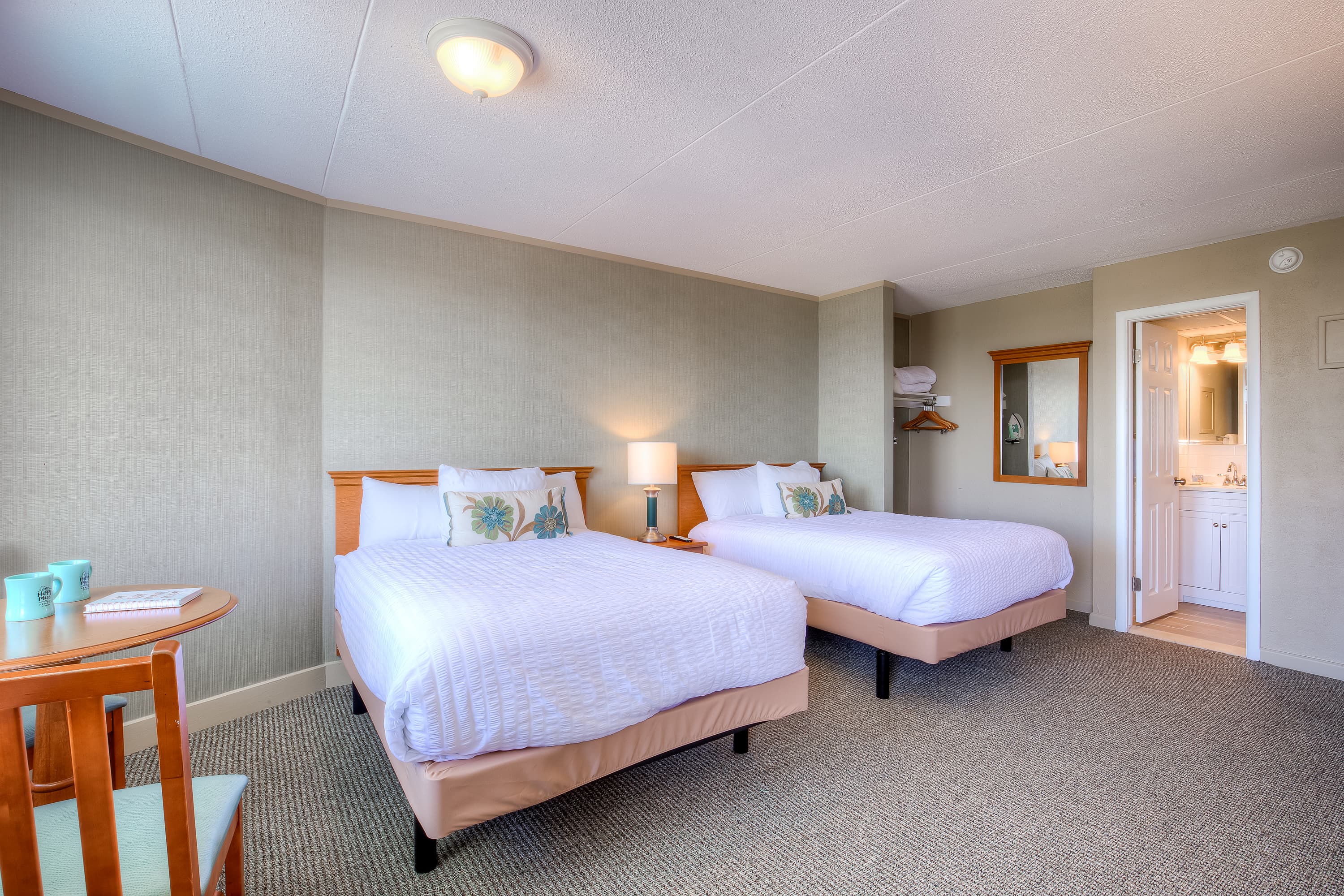 A clean hotel room featuring two double beds, a small table, and a private bathroom.