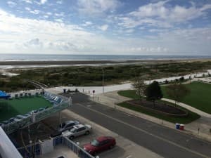 A scenic view of a beach with a cloudy sky, greenery, and parked cars nearby.