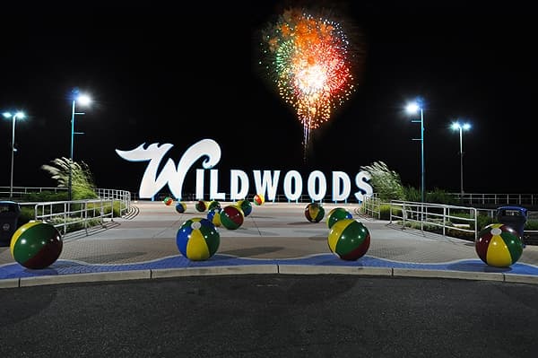 Colorful beach balls in front of the illuminated "Wildwoods" sign under fireworks at night.