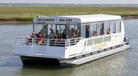 A group of people waves from a tour boat named "The Skimmer" as it navigates through a marsh.