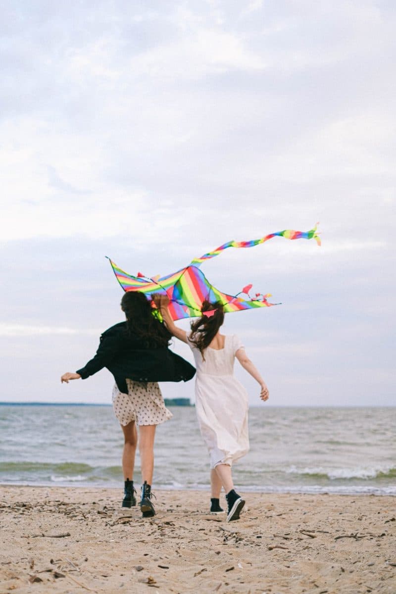 Two girls run along the beach, joyfully holding a rainbow-colored kite.