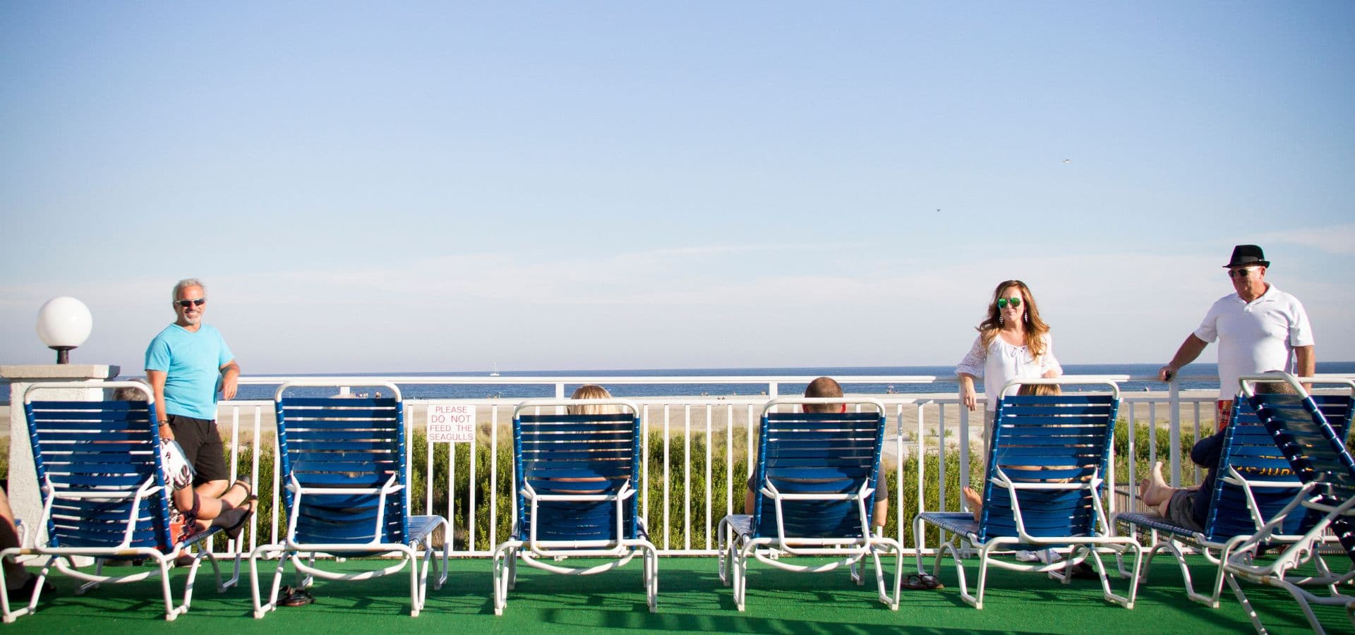 A group of people relaxes on blue lounge chairs by the ocean under a clear sky.