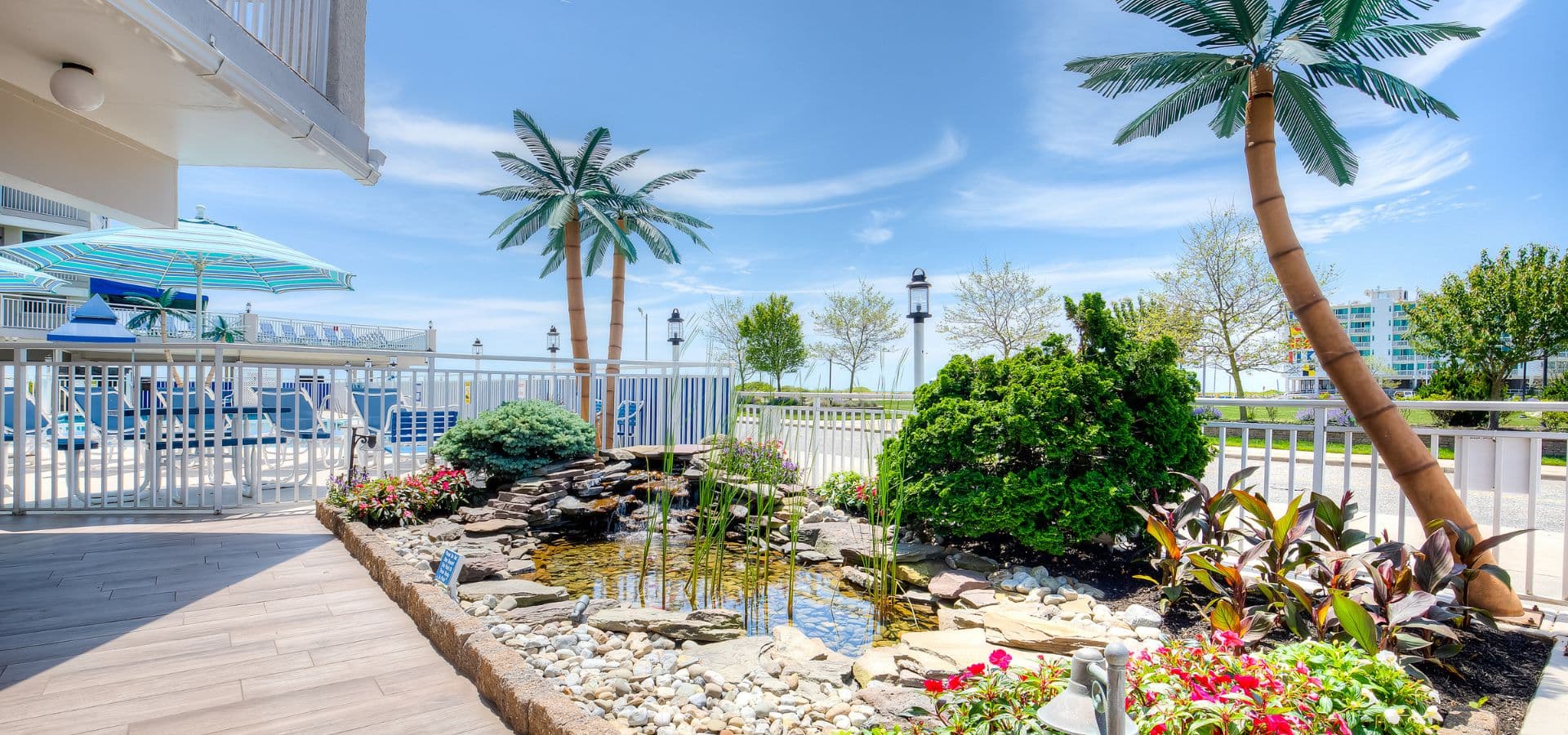 A serene outdoor space featuring a pond surrounded by lush greenery and decorative palm trees under a bright blue sky.