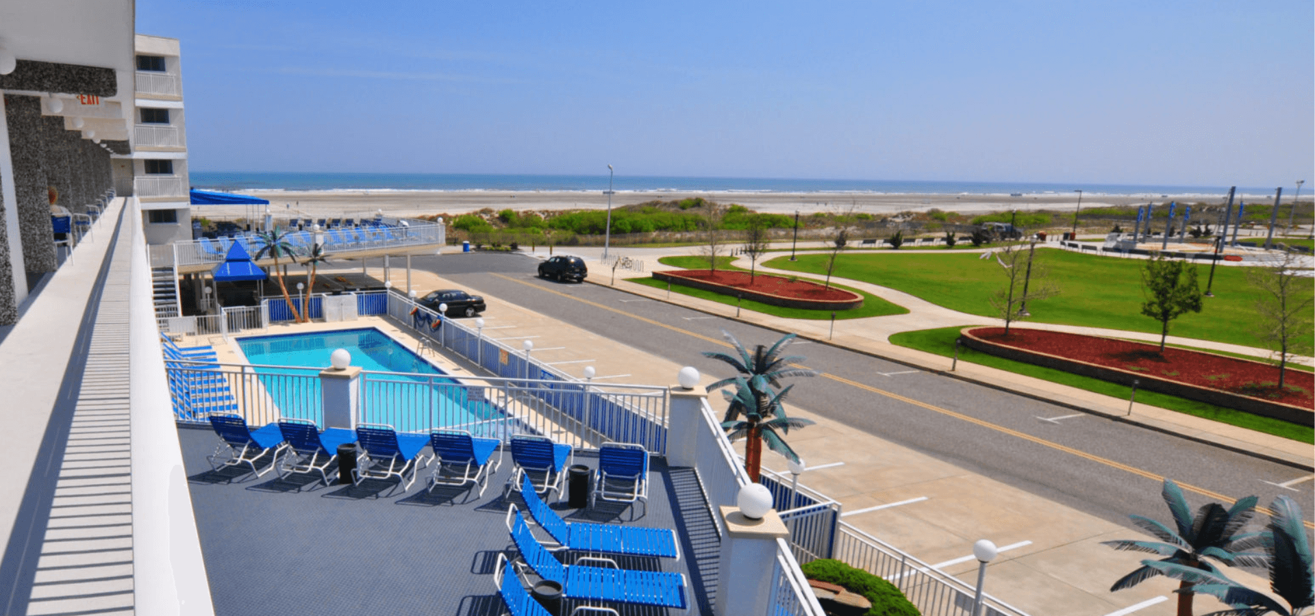 A view of a beachfront hotel pool area overlooking the ocean and a landscaped park.