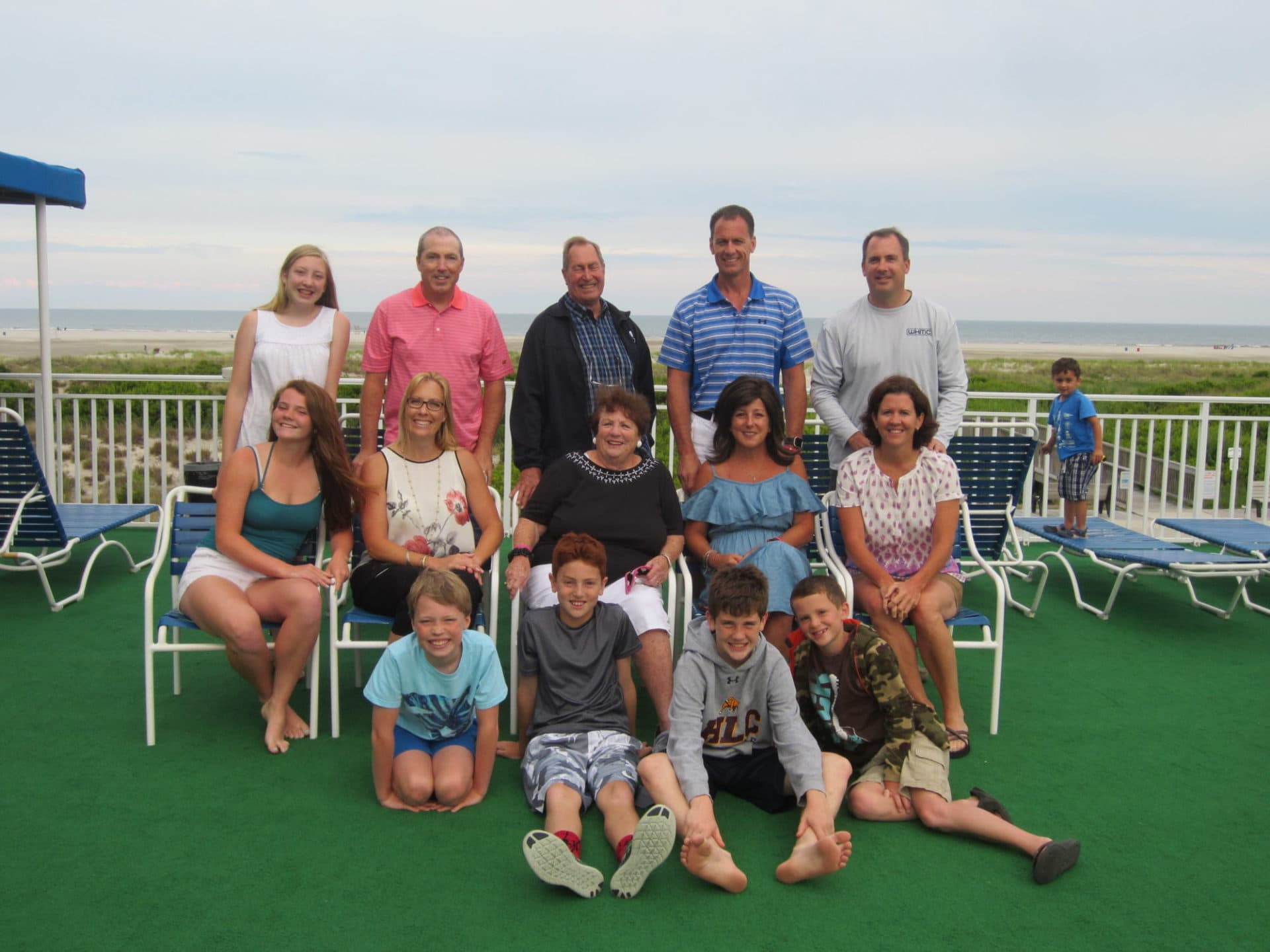 A large family poses for a group photo on a green patio with a beach view in the background.
