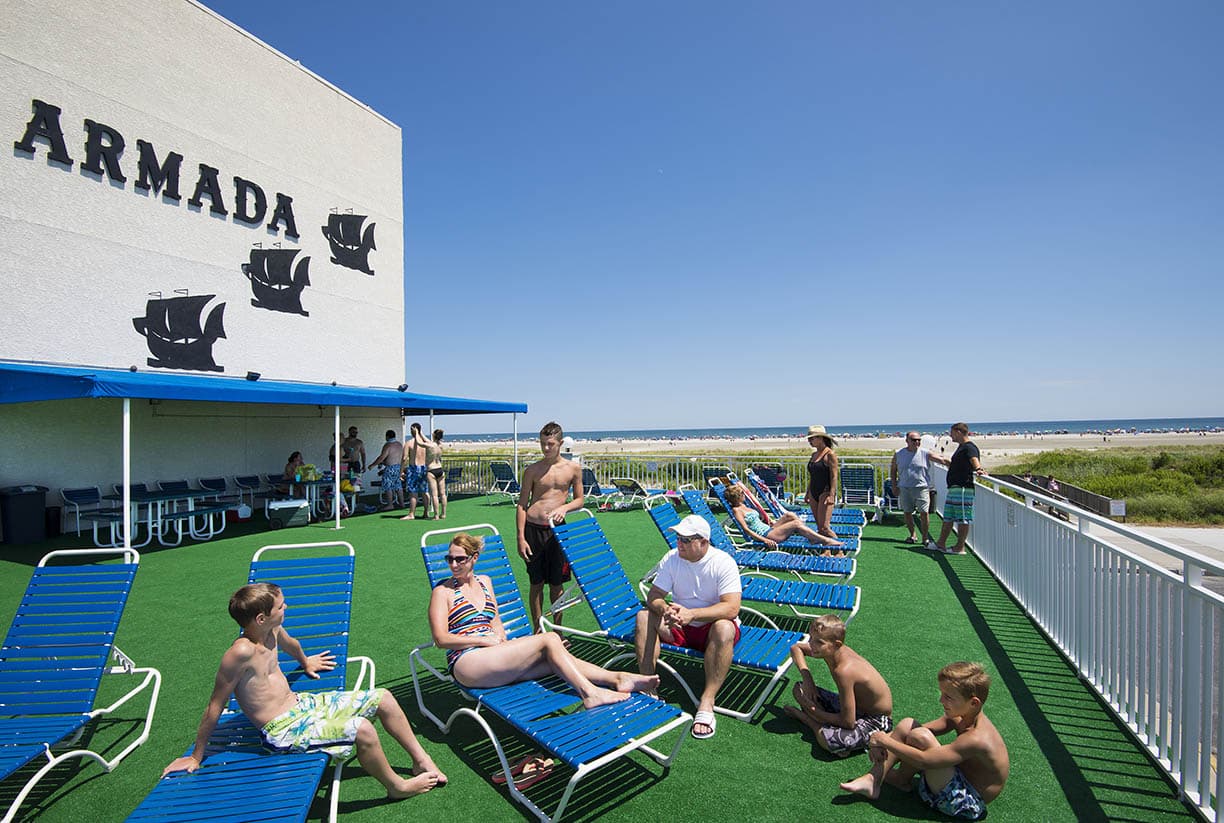 A group of people relaxes on blue lounge chairs by the beachside, under a clear sky and next to a building labeled "Armada."
