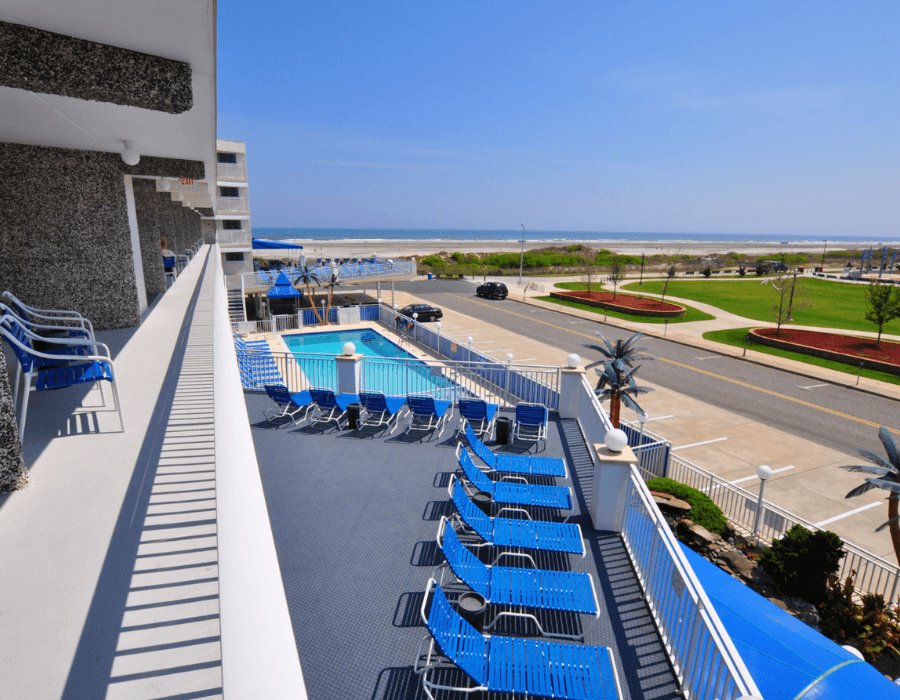 View from a balcony overlooking a pool area with blue lounge chairs and a beach in the distance.