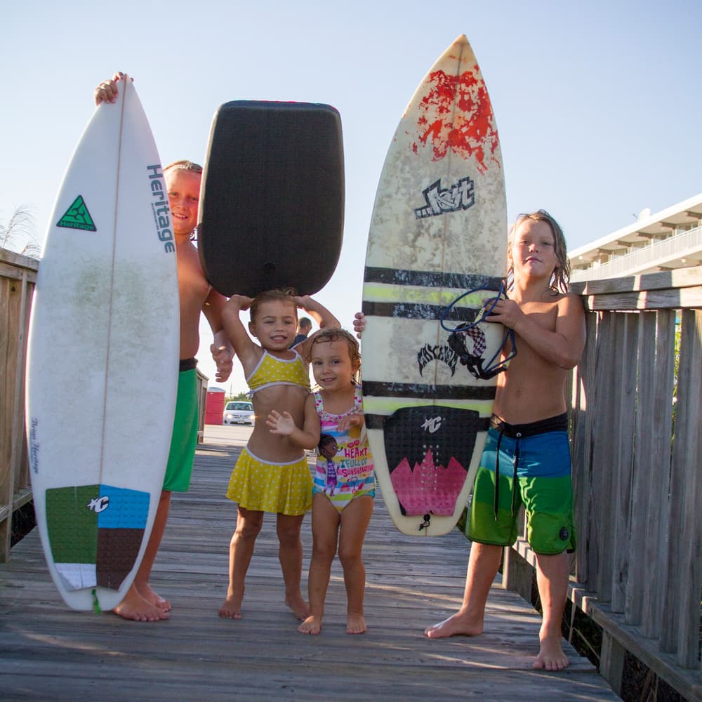 Three children hold surfboards on a wooden dock with a bright blue sky in the background.