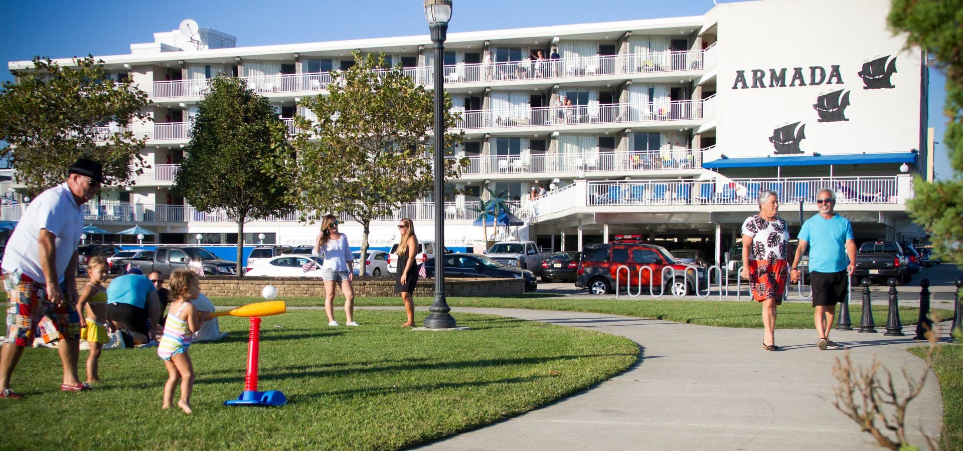 A family plays in a grassy area near the Armada hotel, with people walking and cars parked in the background.