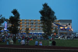 A large crowd gathers in front of a hotel in the evening, surrounded by trees and outdoor seating.