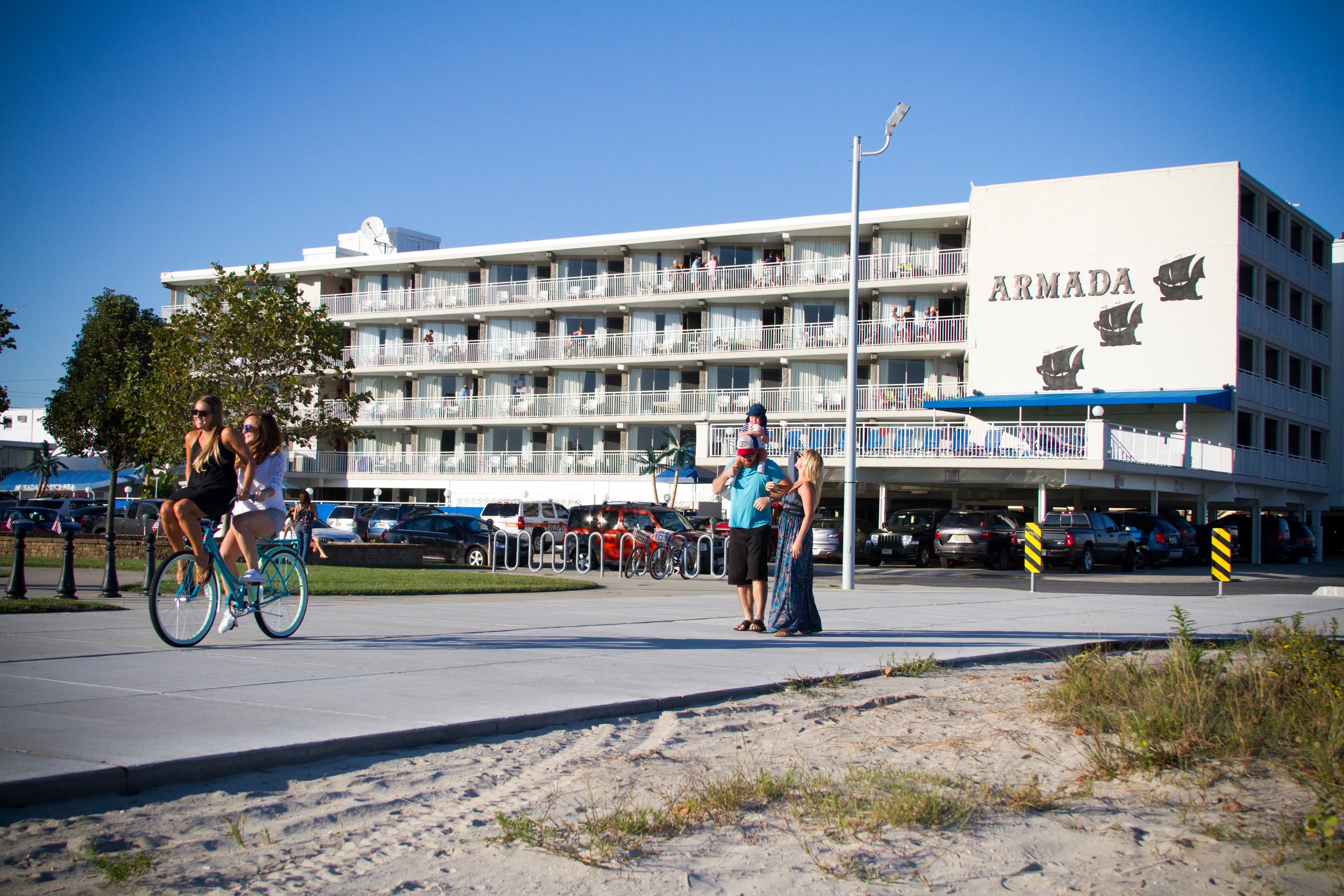 Two women ride bicycles past the Armada hotel while other visitors gather nearby.