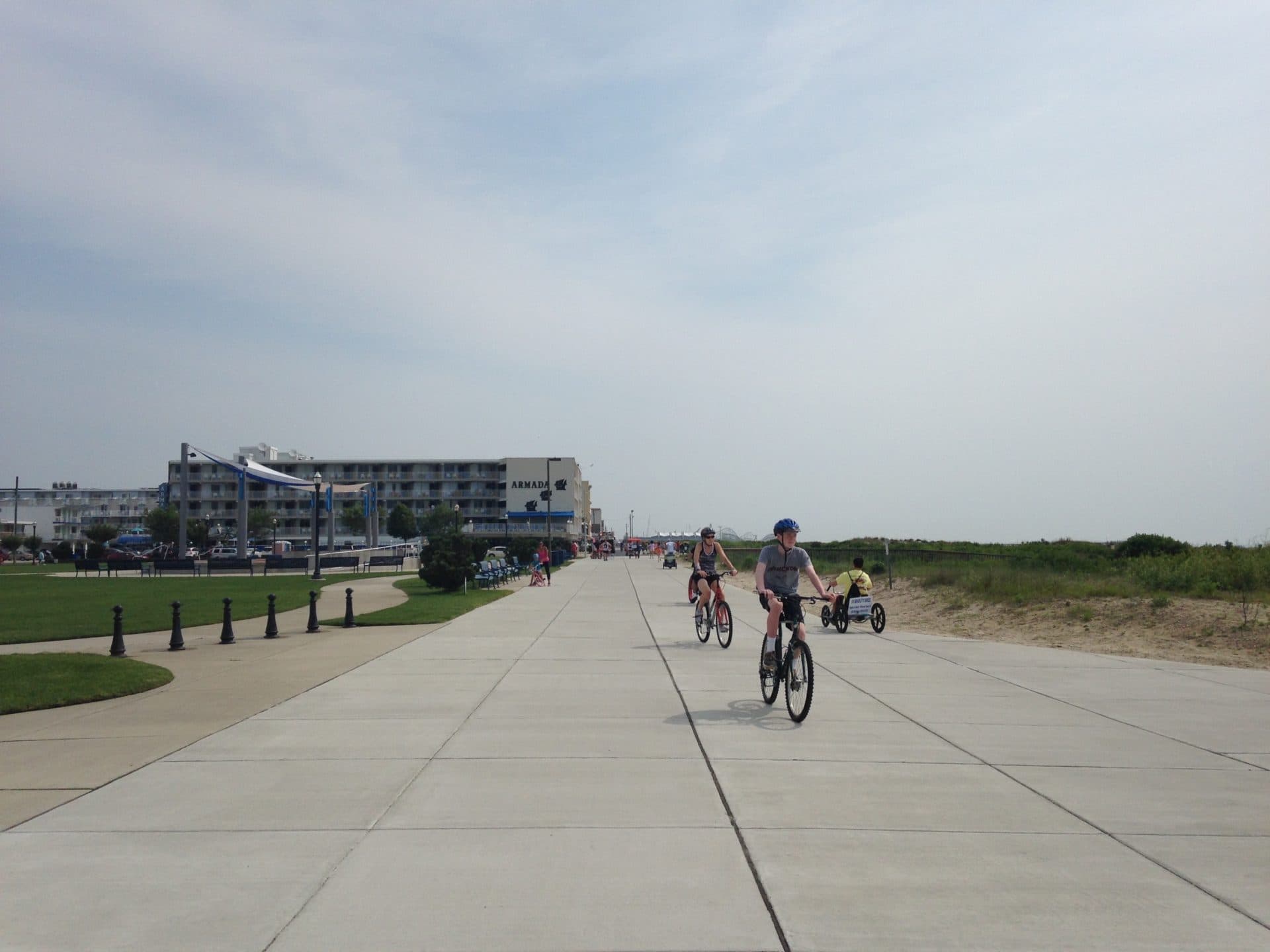 Cyclists ride along a paved path with a beachfront hotel in the background under a cloudy sky.