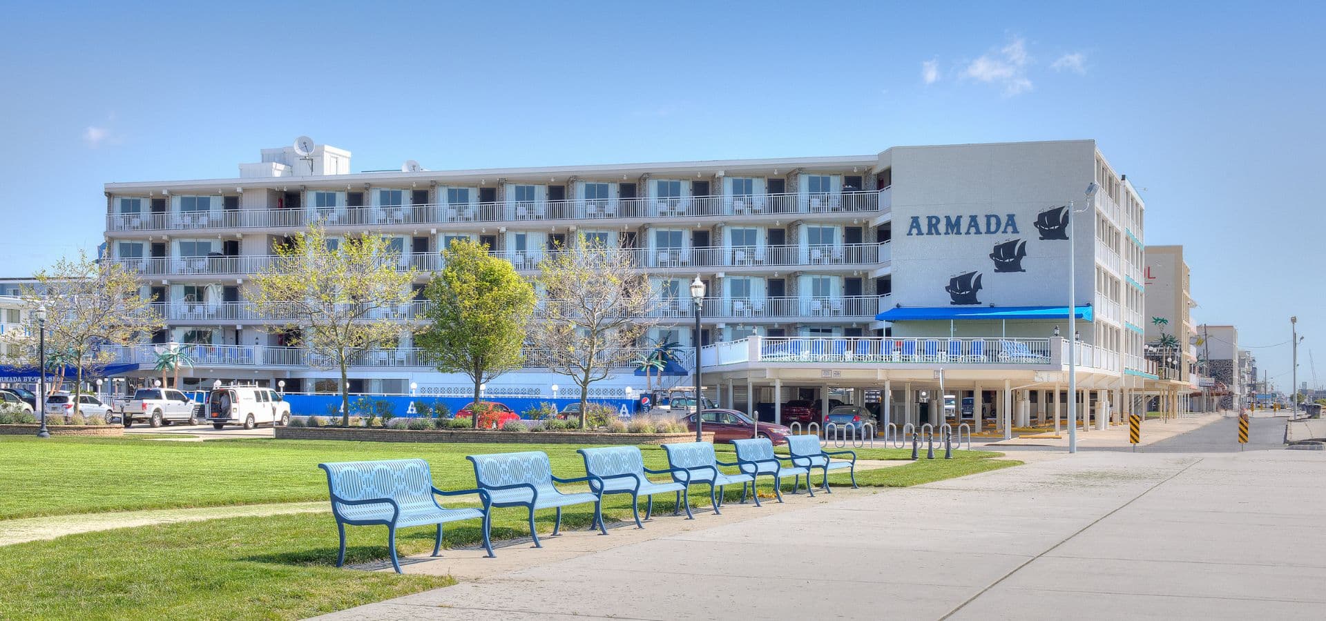 A beachfront hotel named Armada with several benches in front and greenery around it.